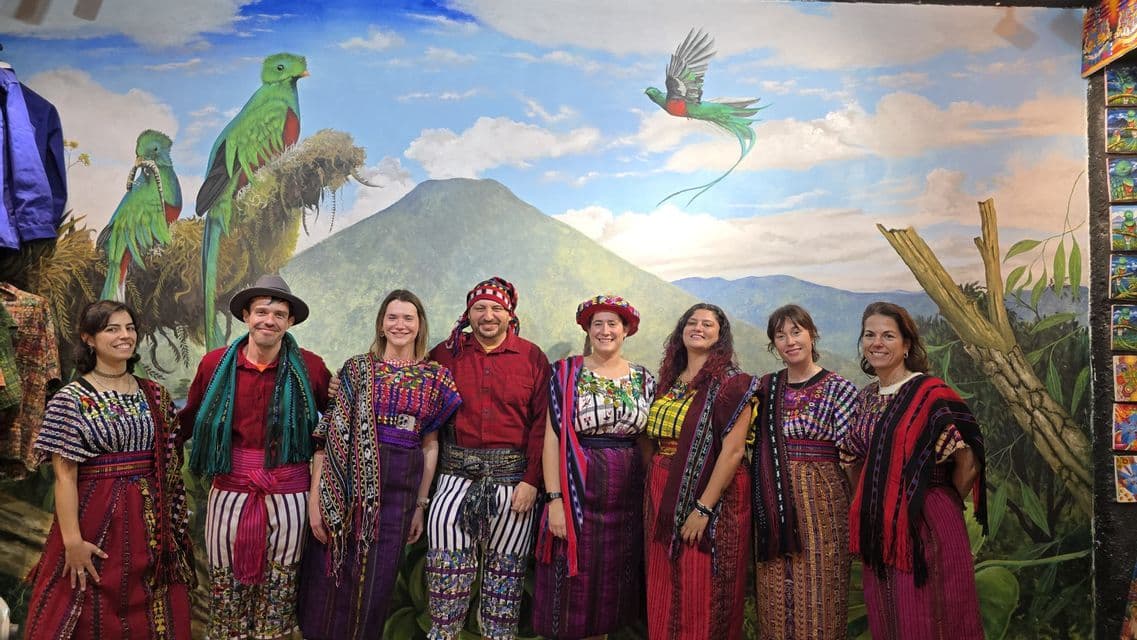 Un viaje en grupo de WeRoad posando con ropa tradicional colorida frente a un gran mural de un volcán y aves quetzal verdes.