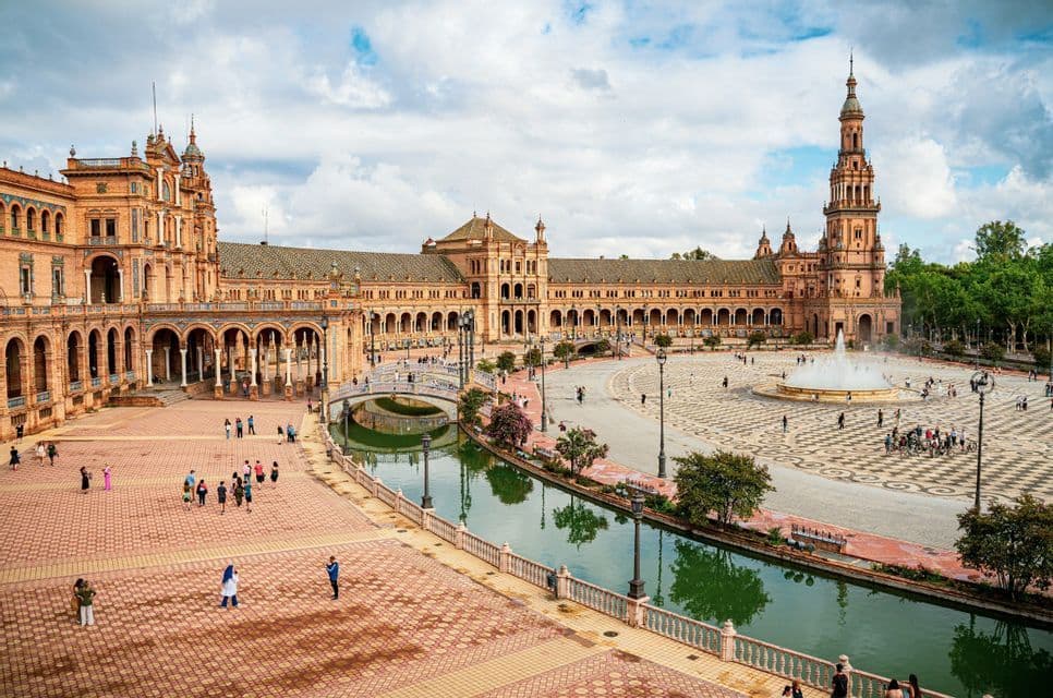 Une vue surélevée d'une grande place avec un bâtiment orné et arcadé, un canal et des gens qui se promènent sous un ciel nuageux.