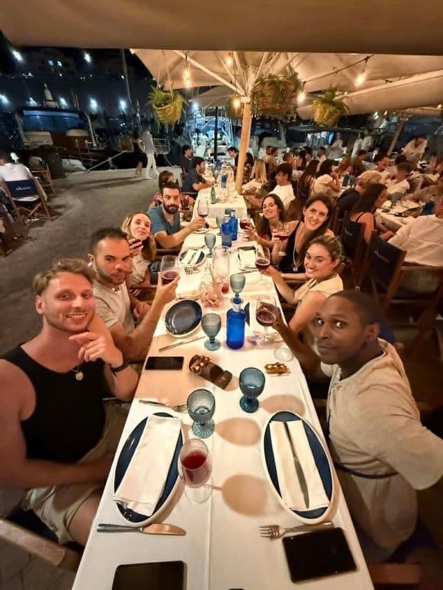 Un groupe WeRoad en voyage dîne à une longue table dans un restaurant en plein air la nuit, souriants et levant leurs verres de vin.