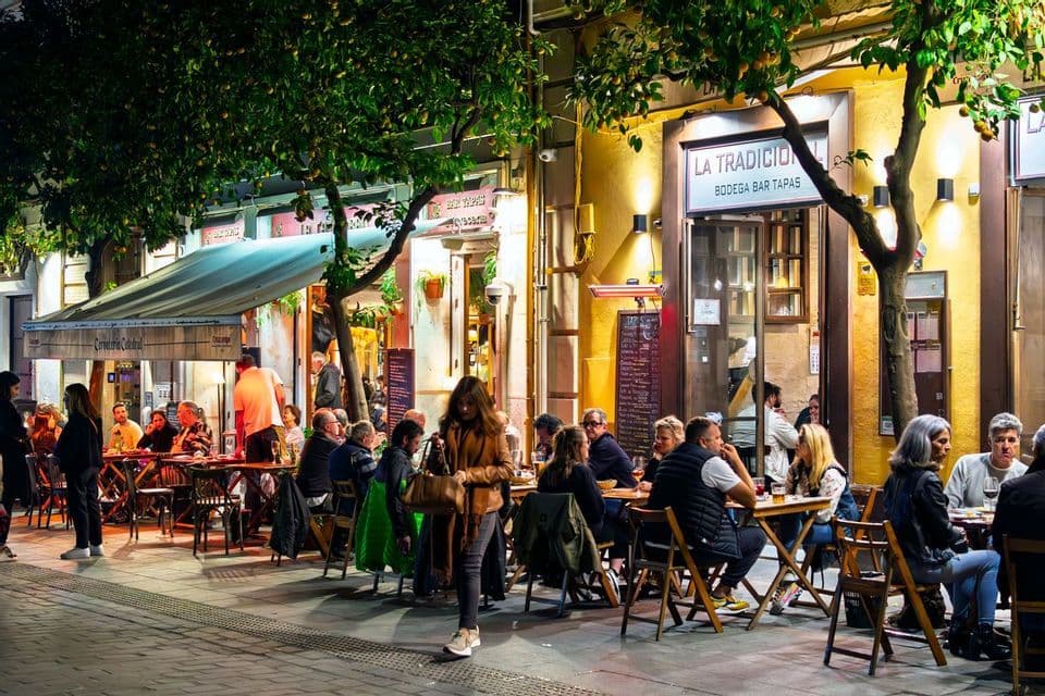 Ambiance nocturne dans une rue pavée : des gens dînent et socialisent en terrasse d'un bar à tapas.