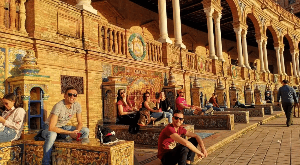 Un groupe WeRoad en voyage se détend sur des bancs ornés et carrelés, dans une place ensoleillée aux arcades en briques.