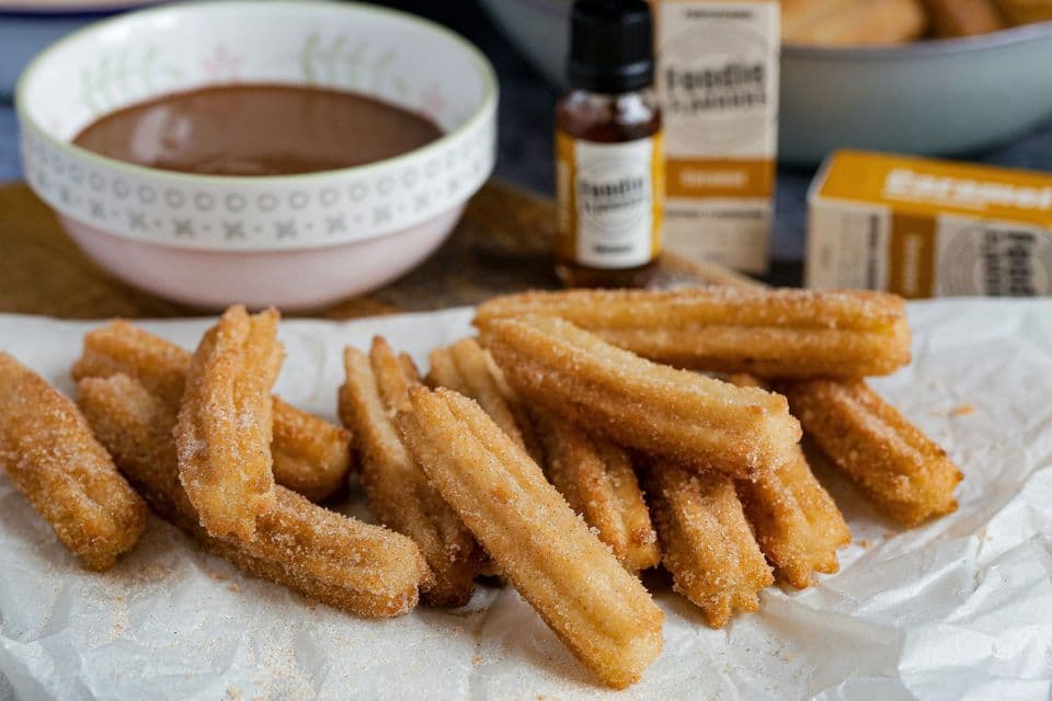 Une pile de churros saupoudrés de sucre sur du papier blanc, avec un bol de sauce au chocolat en arrière-plan.