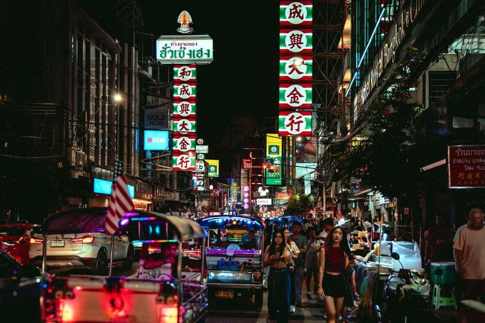 Una vivace strada cittadina di notte, affollata di persone e tuk-tuk, illuminata da grandi insegne al neon colorate.