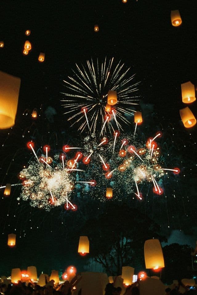 Fuochi d'artificio esplodono in un cielo notturno buio, con numerose lanterne di carta illuminate che si librano verso l'alto da una folla sottostante.