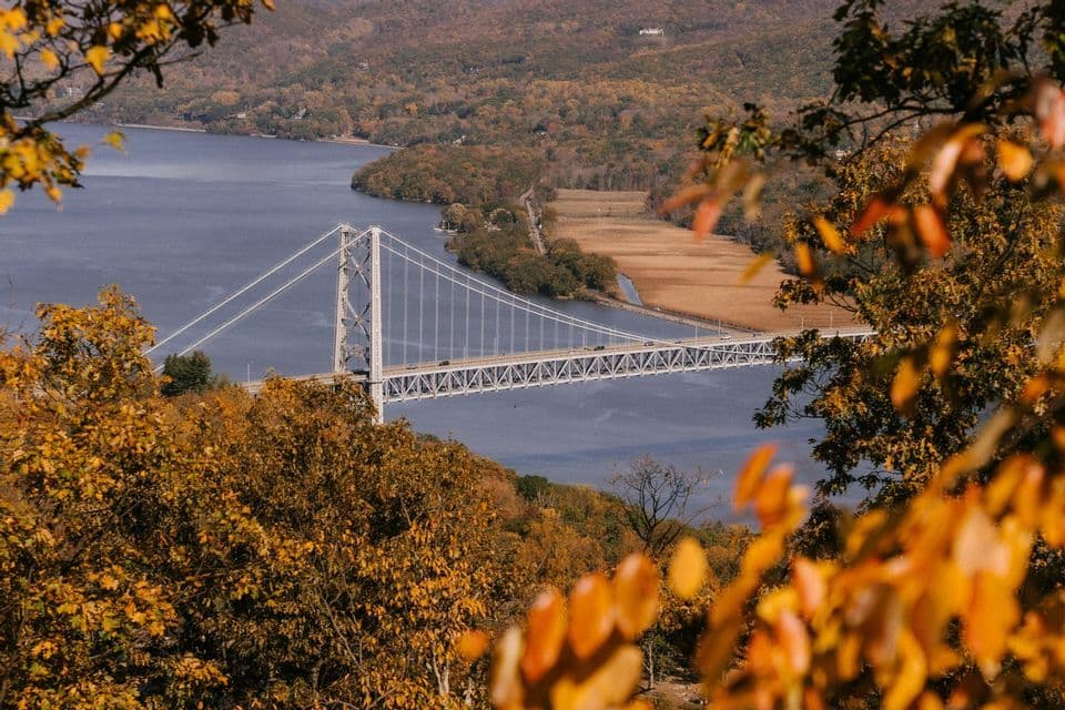 A white suspension bridge spans a river, framed by golden autumn leaves and hills with fall foliage.