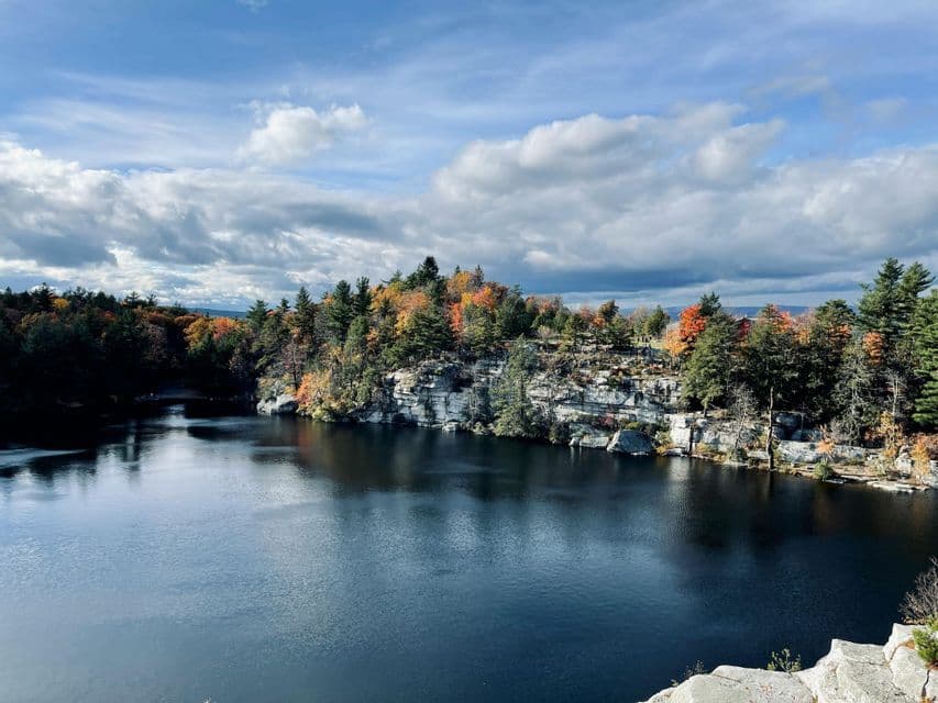 A calm lake surrounded by rocky cliffs and a forest with colorful autumn foliage under a cloudy sky.