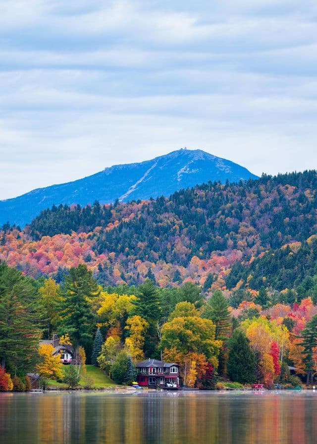 A lakeside house is surrounded by colorful autumn trees with a large mountain in the background under a cloudy sky.