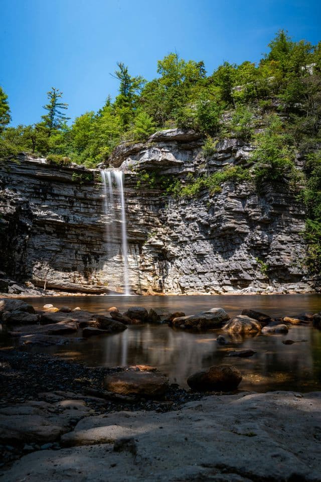 A long-exposure photograph of a waterfall cascading down a layered rock cliff into a calm pool, with trees above against a blue sky.