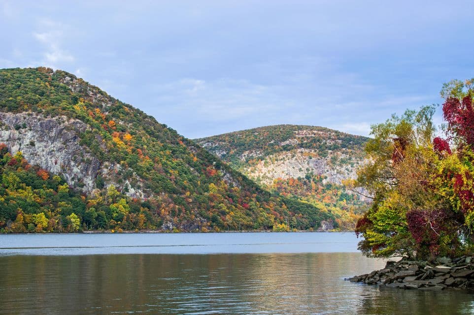 Rolling hills covered in colorful autumn foliage border a wide, calm river under an overcast sky.