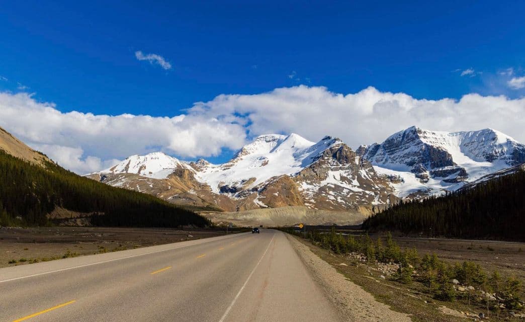 A paved road runs through a valley towards a large, snow-capped mountain range under a blue sky with scattered clouds.