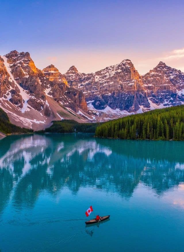 A person in a canoe holding a Canadian flag paddles across a turquoise lake in front of snow-capped mountains and a forest.