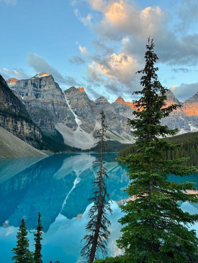 Tall pine trees frame a turquoise alpine lake that perfectly reflects a range of jagged, snow-capped mountains at sunrise.