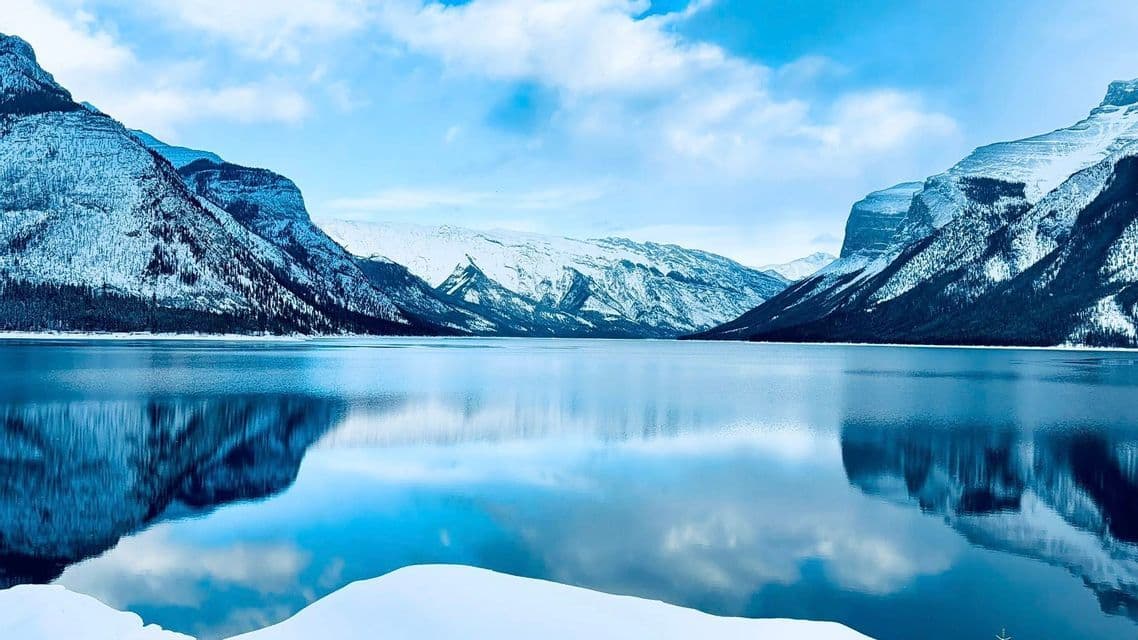 Snowy mountains and a blue sky with clouds reflecting perfectly on the still water of a large lake.