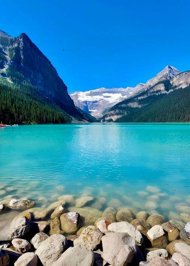 A rocky shoreline on a clear turquoise lake, with pine-covered mountains and a distant glacier under a blue sky.