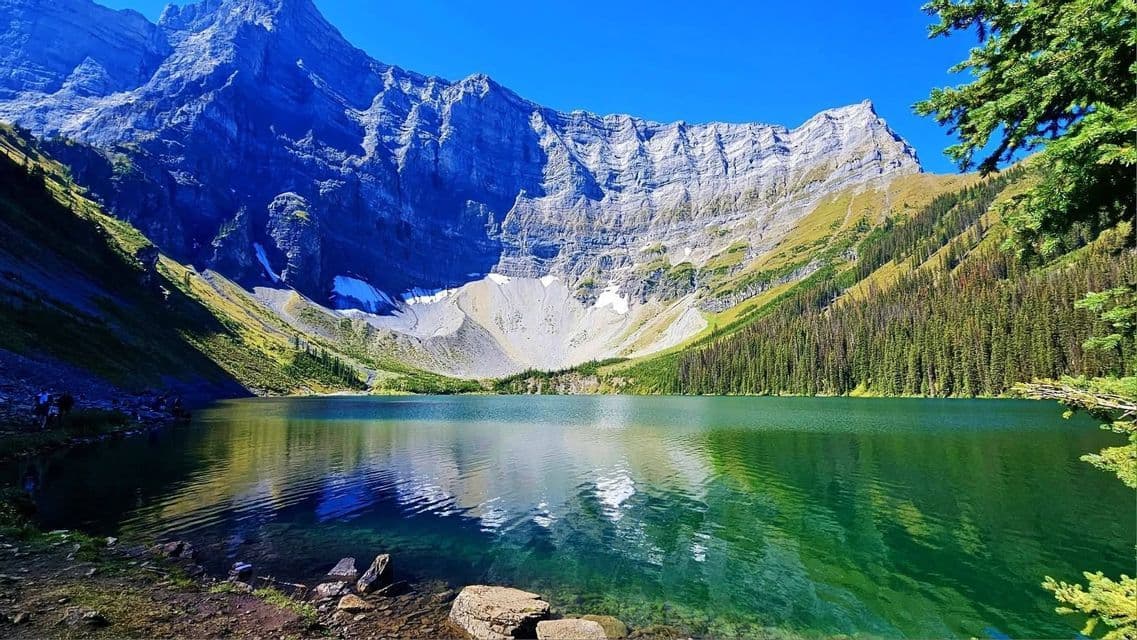 A WeRoad group trip stands by a green alpine lake below a massive rocky mountain cirque under a clear blue sky.