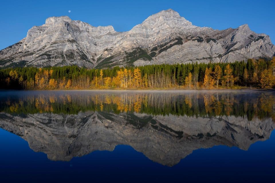 A rocky mountain range and autumn forest perfectly reflected in a still lake under a clear blue sky with a moon.