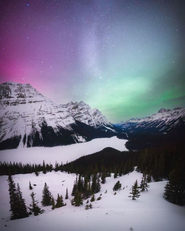 A colorful aurora and starry sky illuminate a snow-covered mountain valley with a frozen lake and pine forest.