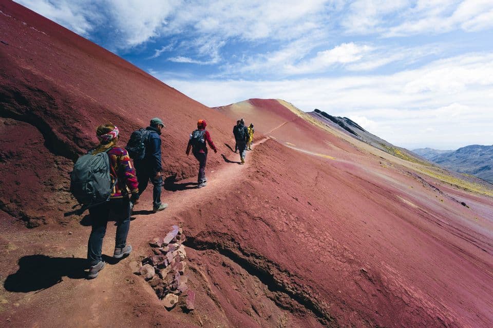 Un groupe WeRoad en randonnée, marchant en file indienne le long d'un sentier étroit à flanc d'une vaste montagne rouge sous un ciel partiellement nuageux.