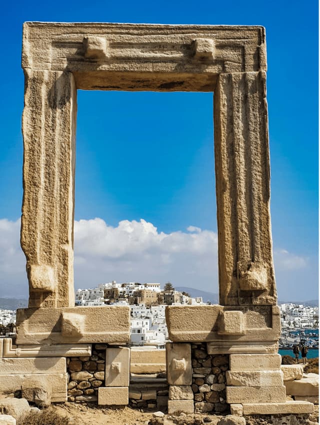Un grande e antico arco in pietra incornicia la vista di un borgo bianco su una collina, sotto un cielo azzurro.