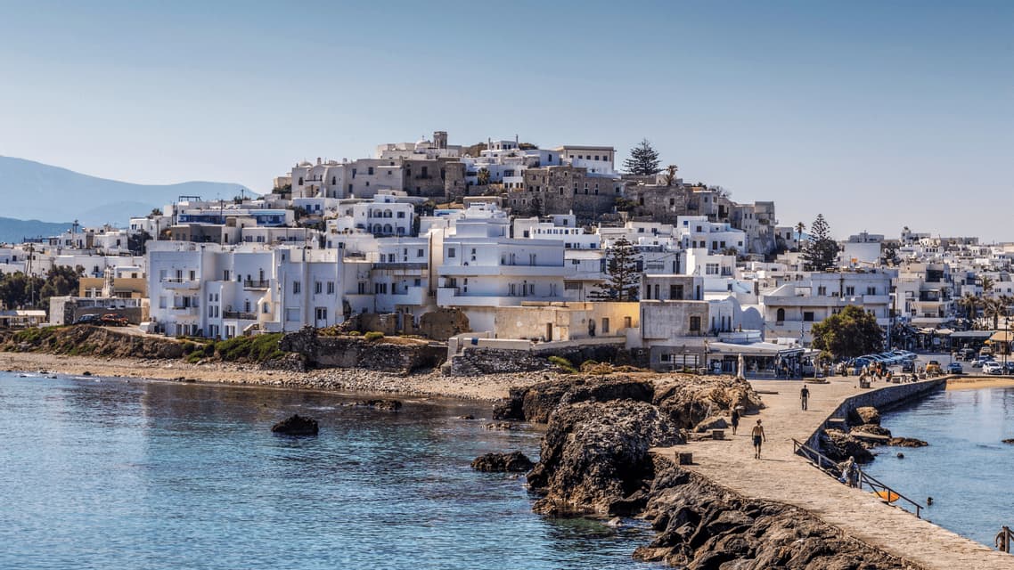 Una cittadina di mare con edifici bianchi su una collina domina un mare azzurro e tranquillo, con un molo in pietra in primo piano.