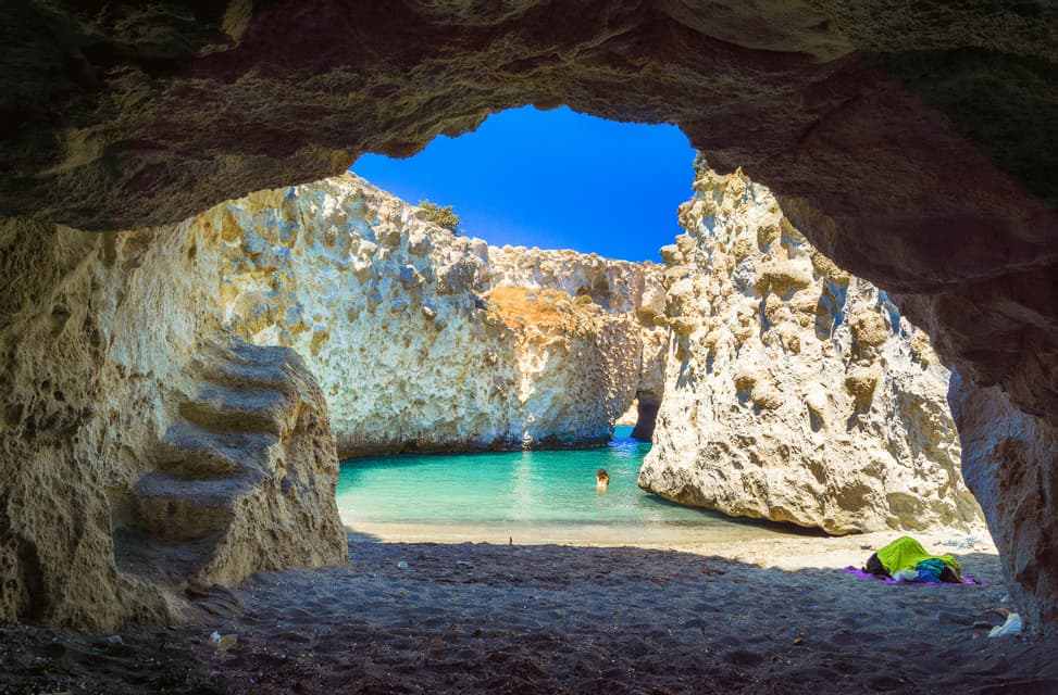 Veduta dall'interno di una grotta marina, con una persona che nuota in una cala turchese circondata da formazioni rocciose bianche.