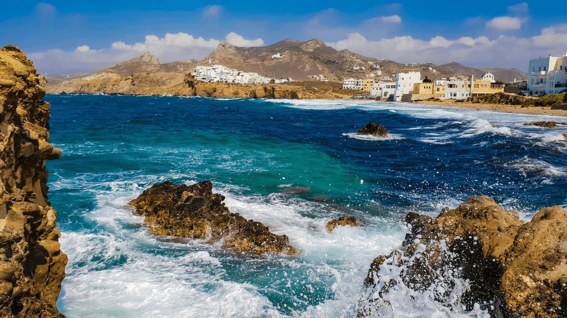 Le onde si infrangono contro una costa rocciosa, con un villaggio costiero di edifici bianchi e montagne sullo sfondo sotto un cielo azzurro.