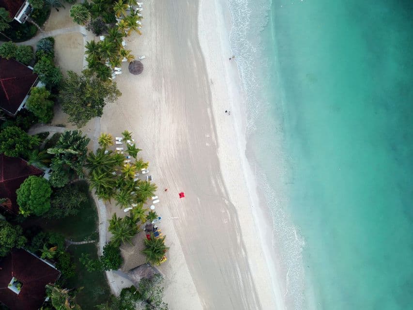An aerial top-down view of a white sand beach bordering turquoise water, with palm trees and resorts on the shore.