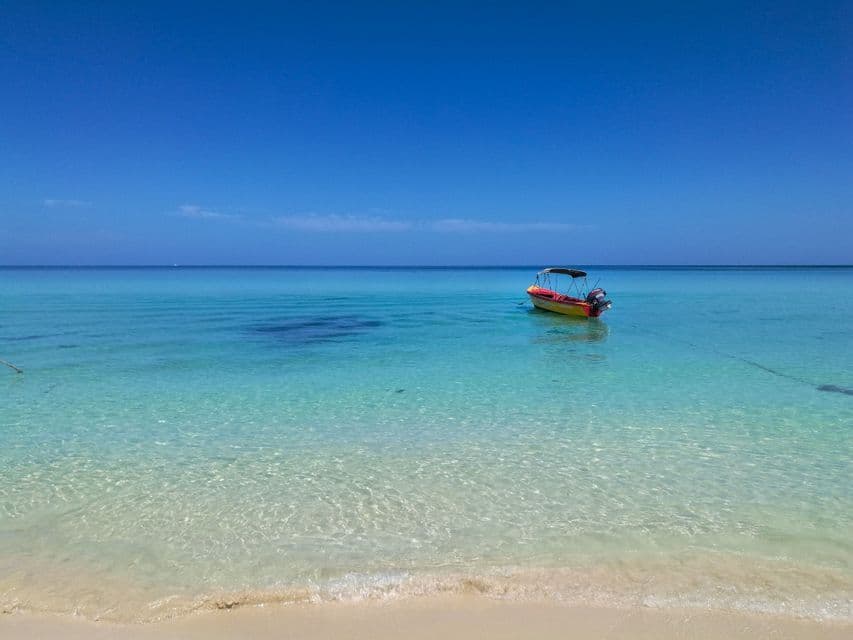 A small yellow and red motorboat is anchored in calm, clear turquoise water off a sandy beach under a deep blue sky.