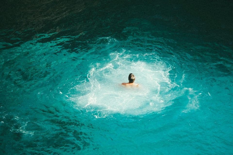 A person seen from behind emerges in the center of a splash in vibrant, turquoise-colored water, as viewed from above.
