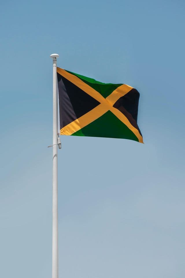 The national flag of Jamaica waving on a white flagpole against a clear blue sky.