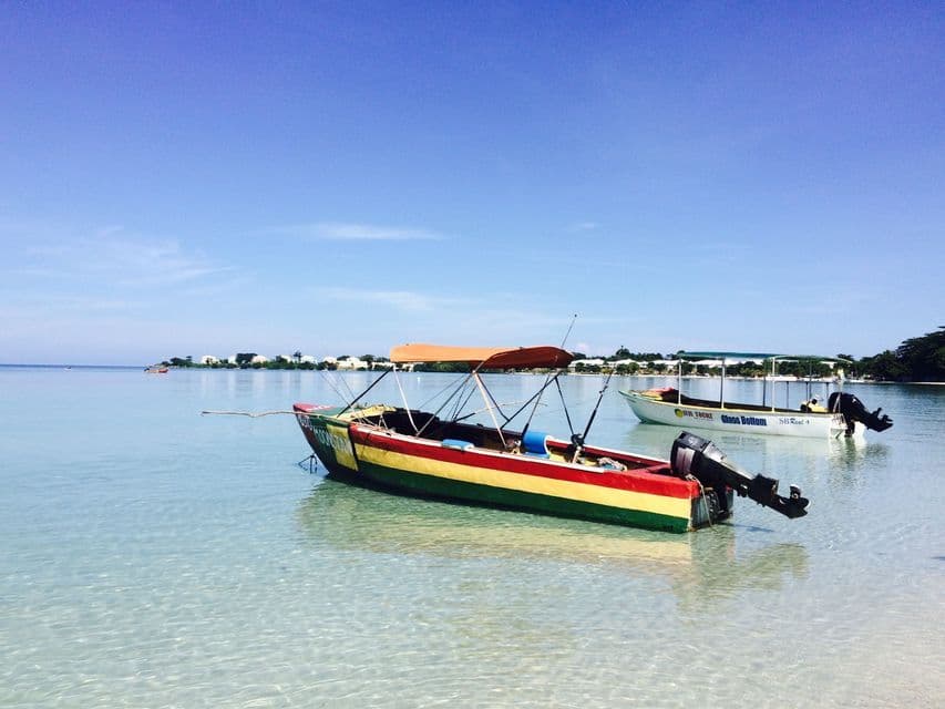 A colorful boat with red, yellow, and green stripes is anchored in shallow, clear turquoise water near a distant shore.