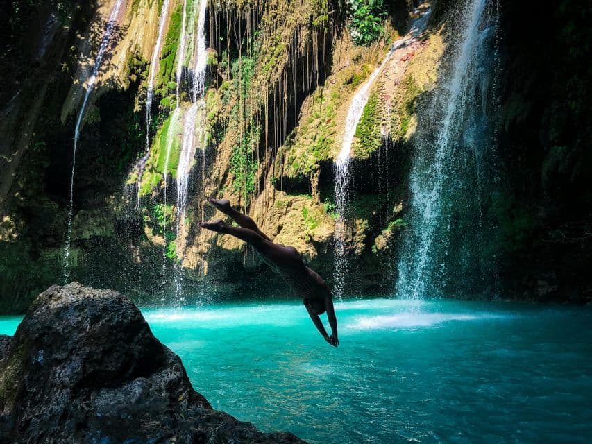 A person dives headfirst from a rock into a turquoise pool at the base of a cascading waterfall.