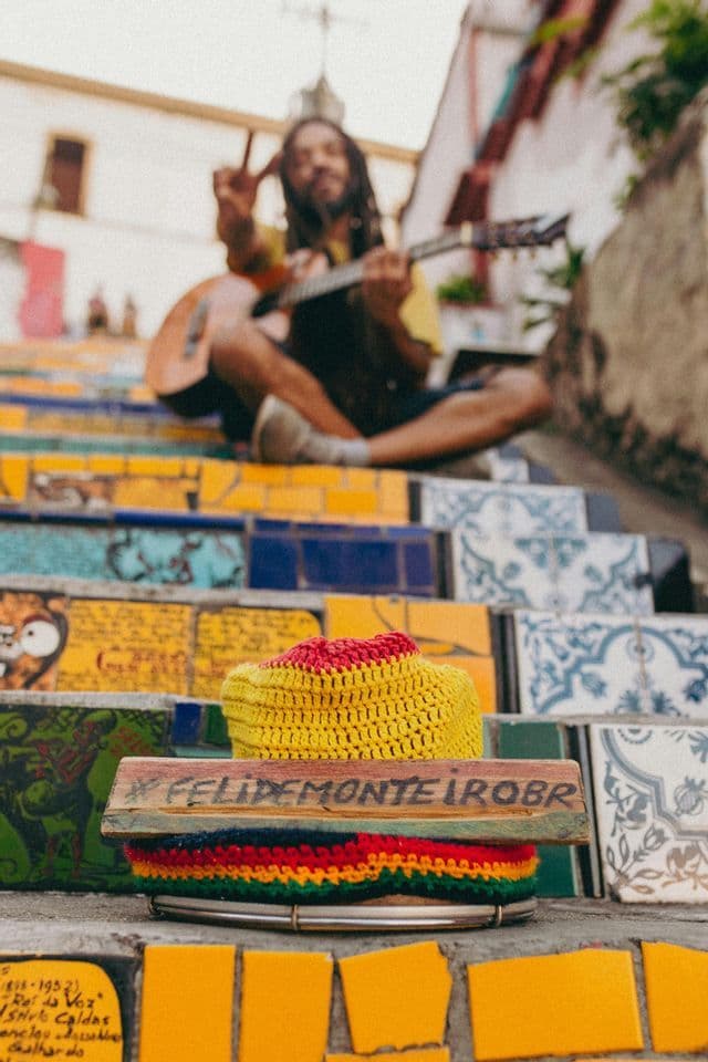 A man with a guitar sits on colorful mosaic stairs making a peace sign, with a crocheted hat in the foreground.