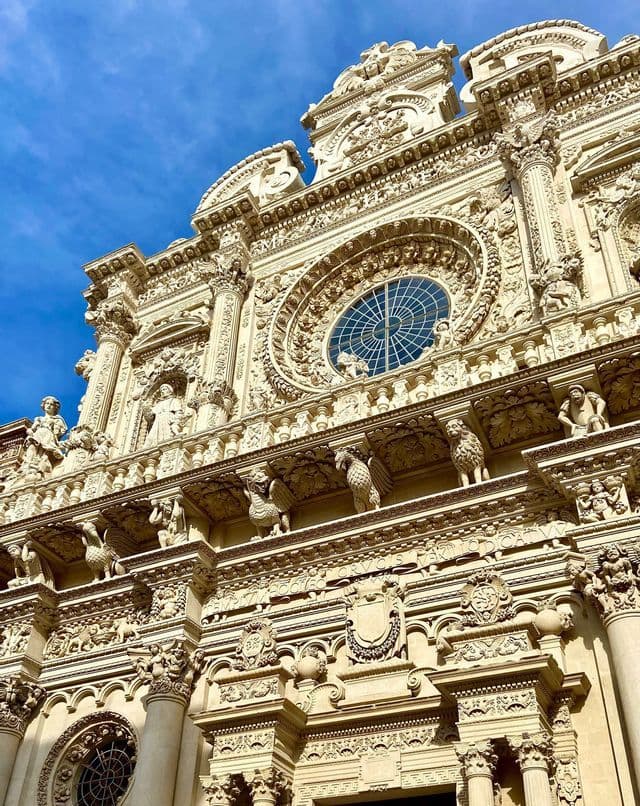 Vista dal basso di una facciata barocca riccamente decorata, con dettagliati intagli in pietra e un grande rosone, contro un cielo azzurro.