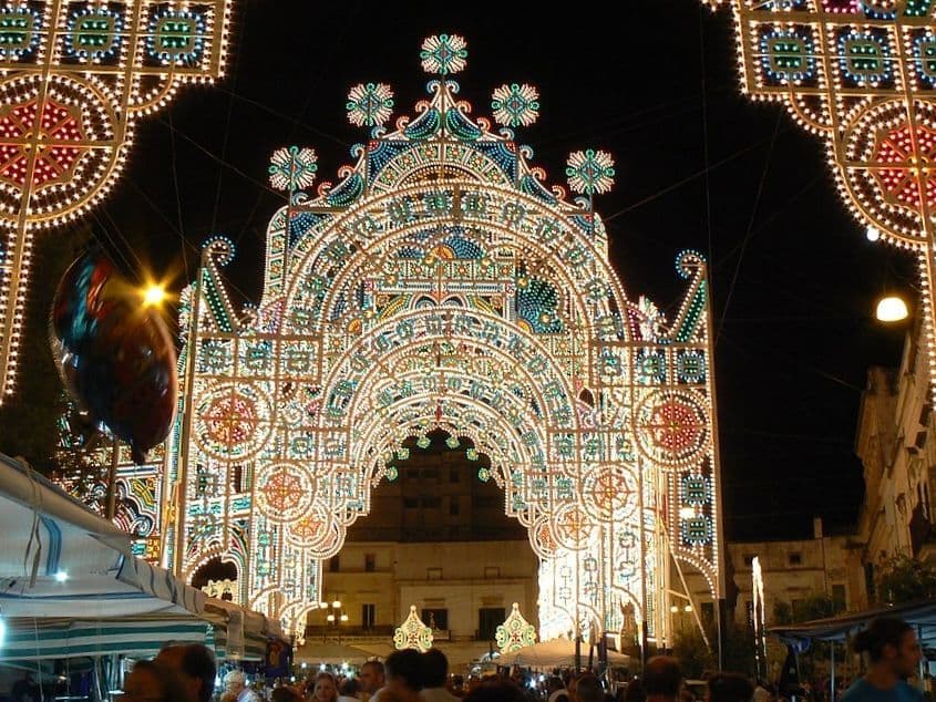 Un sontuoso arco, composto da migliaia di luci colorate, illumina di notte un festival di strada, con una folla di persone sotto.