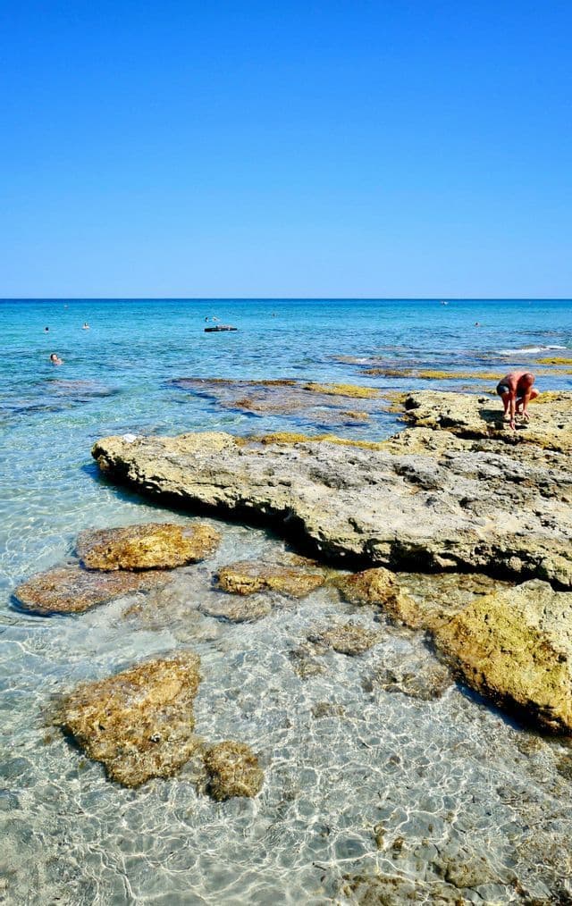 Una persona si china su una riva rocciosa vicino all'acqua limpida e turchese, con gente che nuota in lontananza sotto un cielo azzurro.