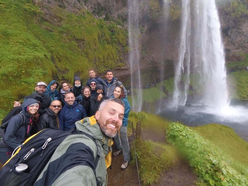 Un gruppo WeRoad sorridente si scatta un selfie su un sentiero, accanto a una grande cascata che scende da una parete rocciosa verde e muschiosa.