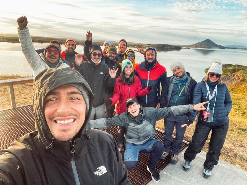 A man takes a selfie with a WeRoad group trip on a viewpoint overlooking a lake and hills under a partly cloudy sky.