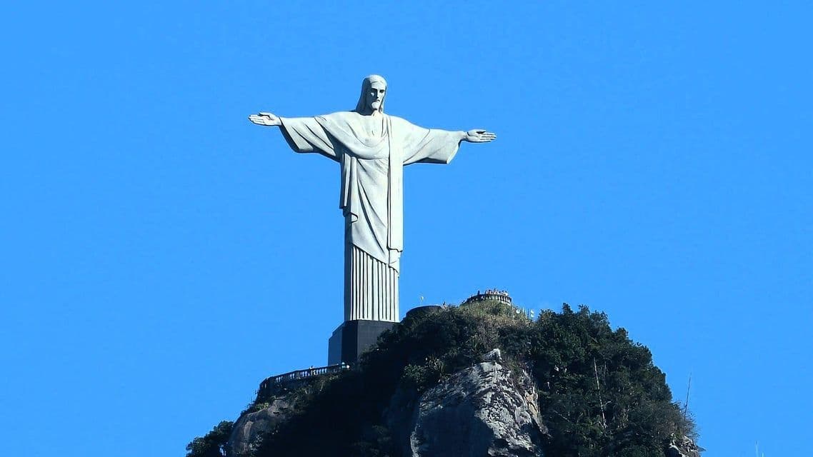 Die Christusstatue steht auf einem felsigen, bewaldeten Berggipfel vor tiefblauem Himmel.