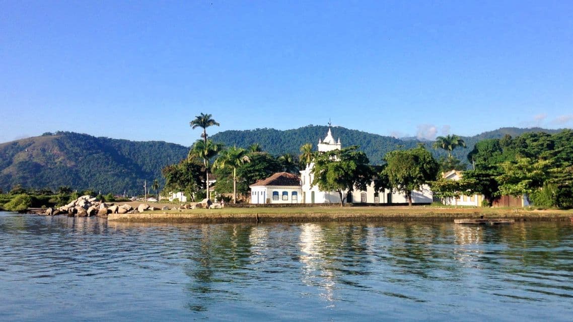 Eine weiße Kirche und Gebäude an einem üppigen, von Bäumen gesäumten Ufer, vom Wasser aus gesehen, mit bewaldeten Bergen im Hintergrund.