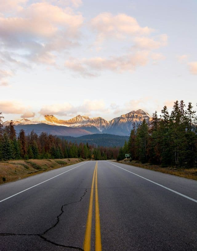 An empty two-lane road with yellow lines leads through a forest towards a distant, sunlit mountain range under a cloudy sky.