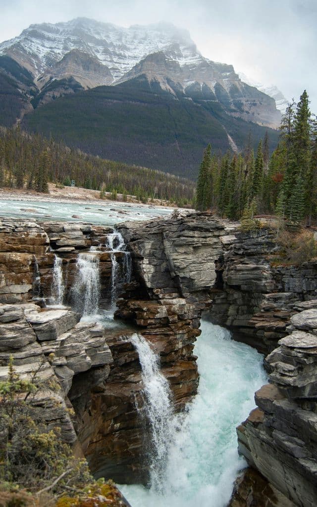 A powerful waterfall cascades through a rocky canyon, with a river and snow-capped mountains in the background.
