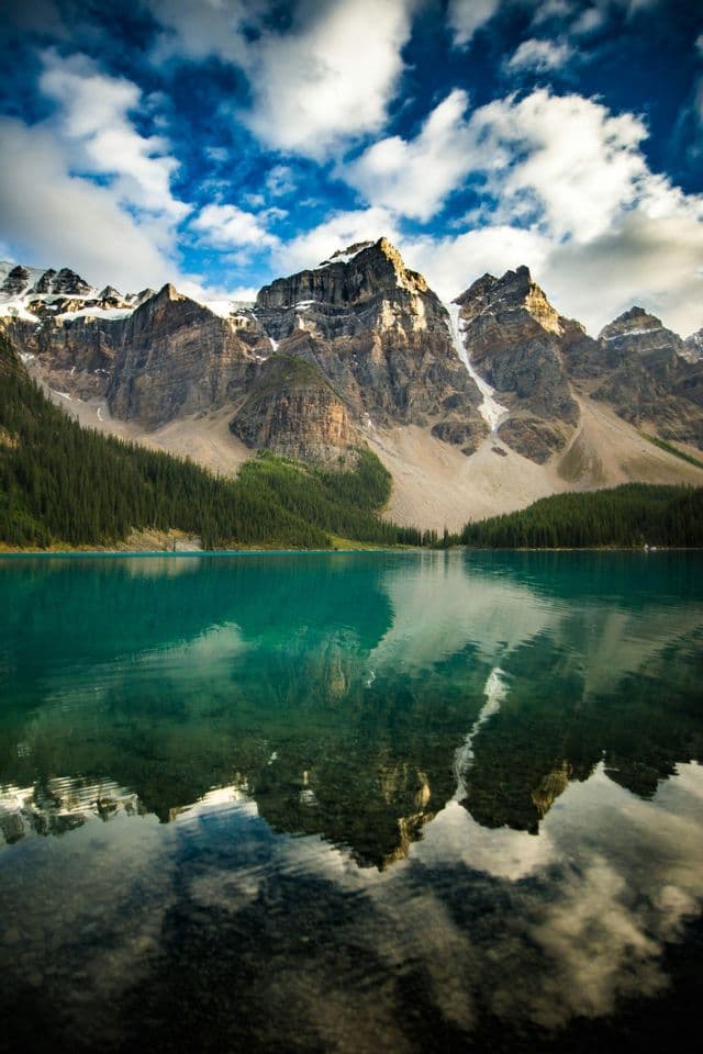 A rocky, snow-capped mountain range is reflected in the still, turquoise water of a lake under a cloudy sky.