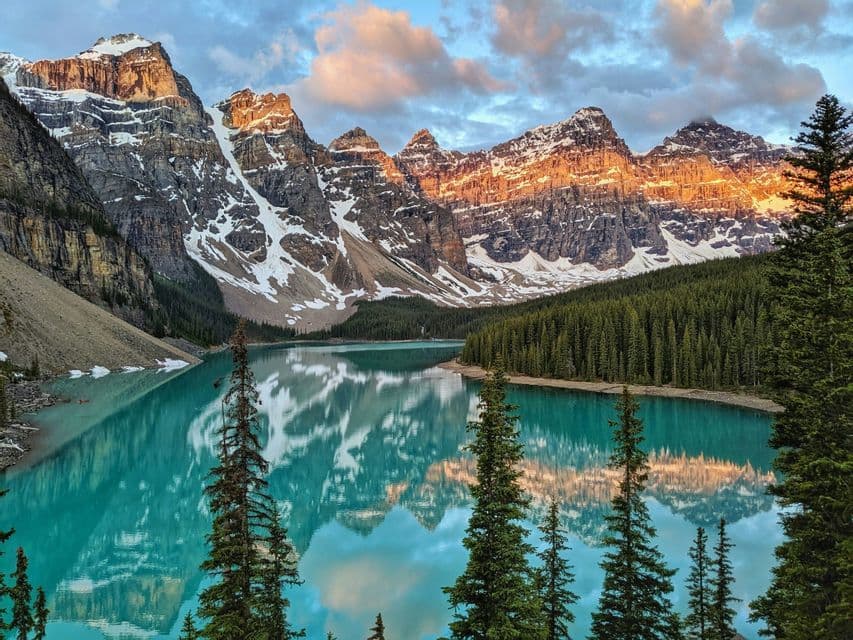 Snow-capped mountains at sunset are reflected in a turquoise lake surrounded by a pine forest.