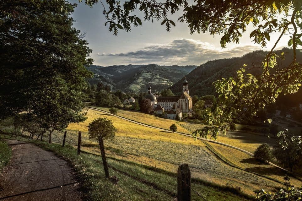 Un monastero storico con cupole si trova in una valle soleggiata, circondato da dolci colline e campi dorati, visibile da un sentiero.