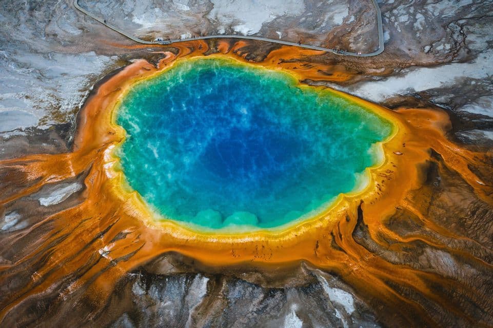 An aerial view of a large geothermal spring with deep blue water surrounded by vibrant orange and yellow mineral deposits.