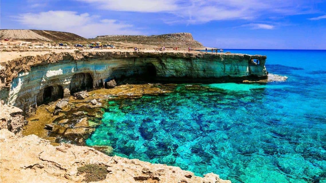 Scogliere rocciose con grotte marine incontrano un mare limpido e turchese, con persone in piedi sulla cima sotto un cielo azzurro.