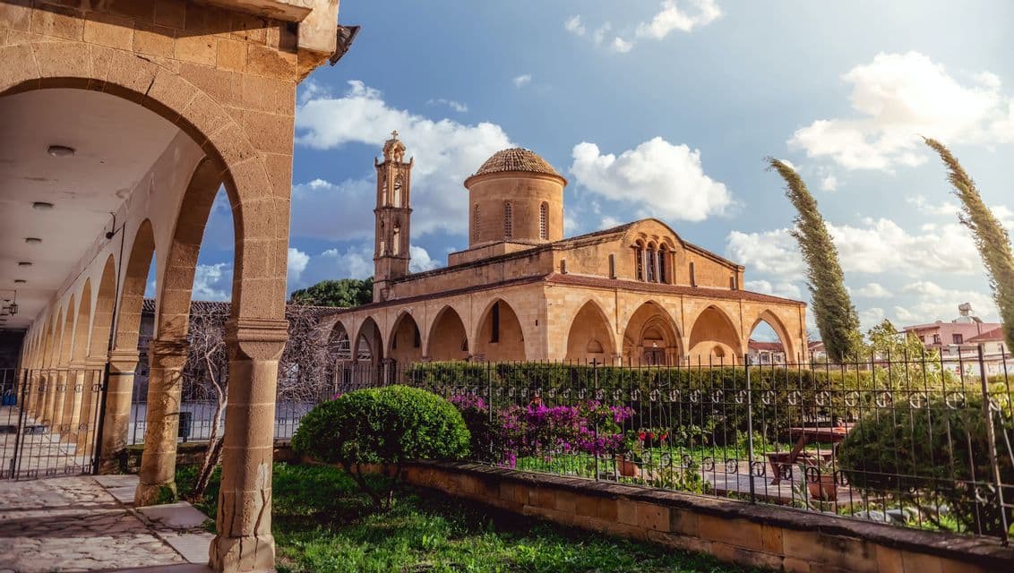 Una chiesa in pietra con cupola e campanile, inquadrata da un arco in pietra, con un giardino e una recinzione in primo piano sotto un cielo azzurro e nuvoloso.