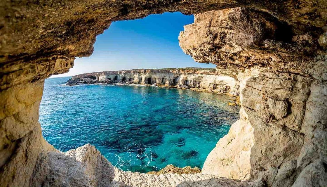 Una vista dall'interno di una grotta marina rocciosa che si affaccia su una costa con scogliere e acqua turchese cristallina sotto un cielo blu.