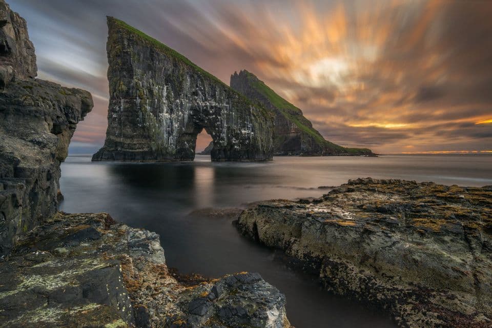 Una foto a lunga esposizione di un grande faraglione con un arco naturale su una costa rocciosa, con nuvole striate durante un tramonto.